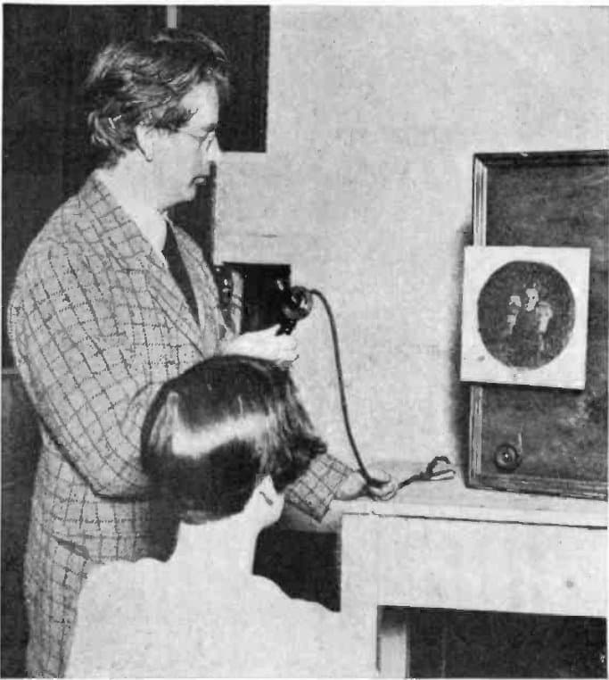 John Logie Baird beside an early television receiver showing a face.